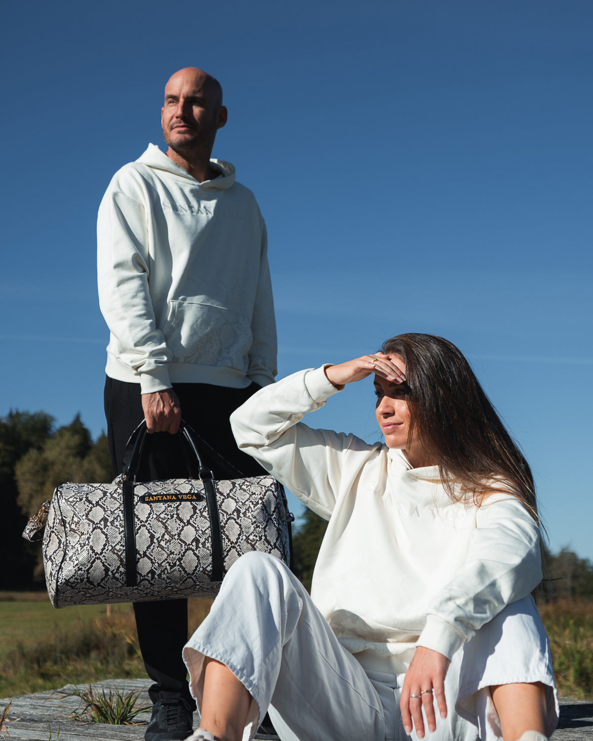 Mann mit Snake Reisetasche und Frau in Off-White Hoodies von Santana Vega unter blauem Himmel