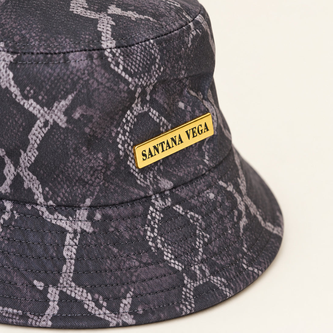 Nahaufnahme der goldenen Plakette auf dem dunkelgrauen Snake Bucket Hat von Santana Vega