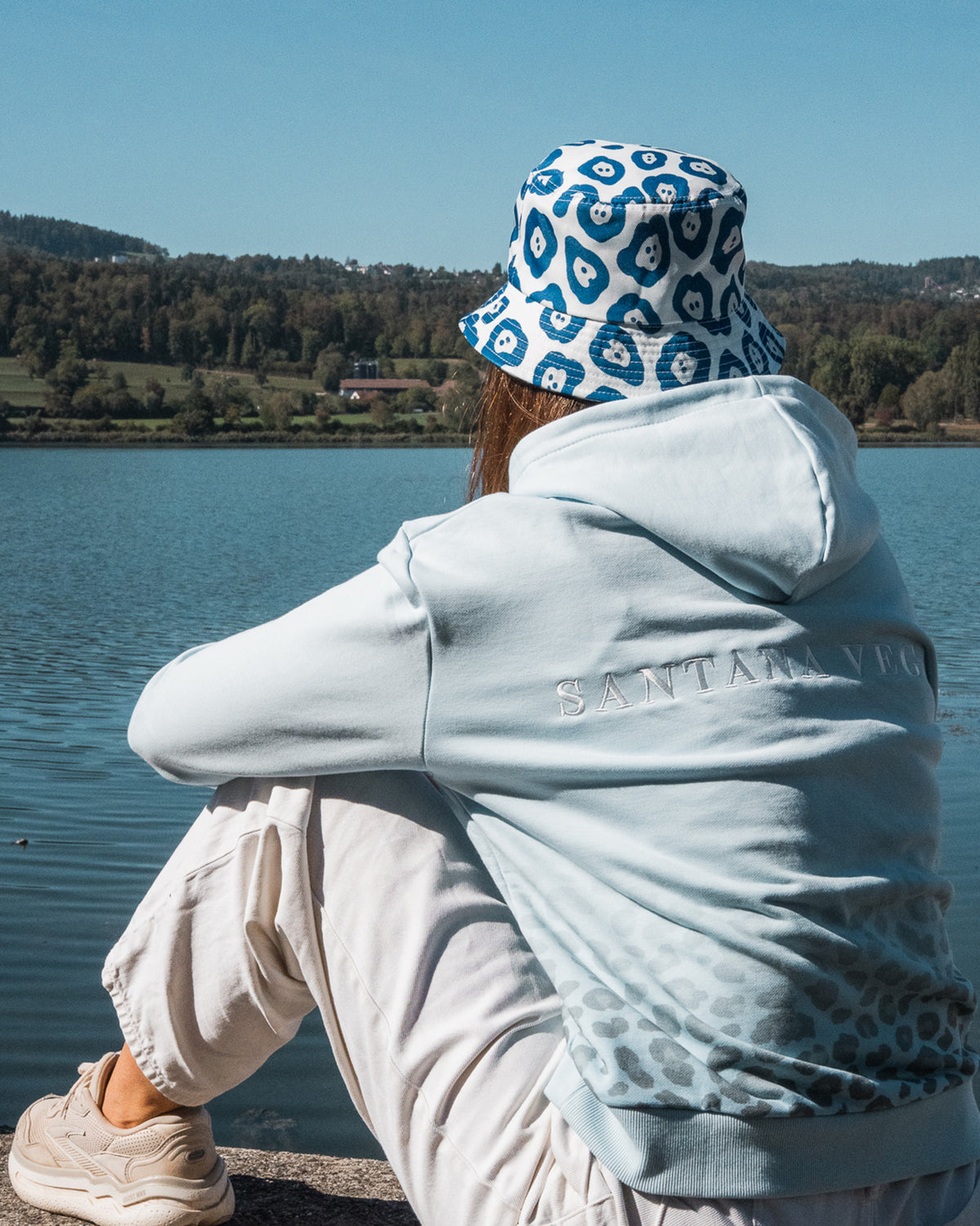 Frau sitzt am See, Rückansicht mit Jaguar Bucket Hat und Santana Vega Stickerei auf dem hellblauen Leopard Hoodie